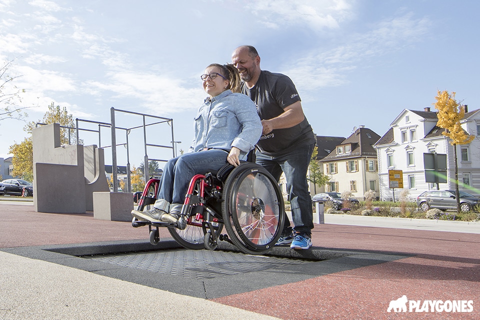 une jeune fille en situation d'handicap sur un fauteuil roulant saute sur un trampoline, aidée par son parent.