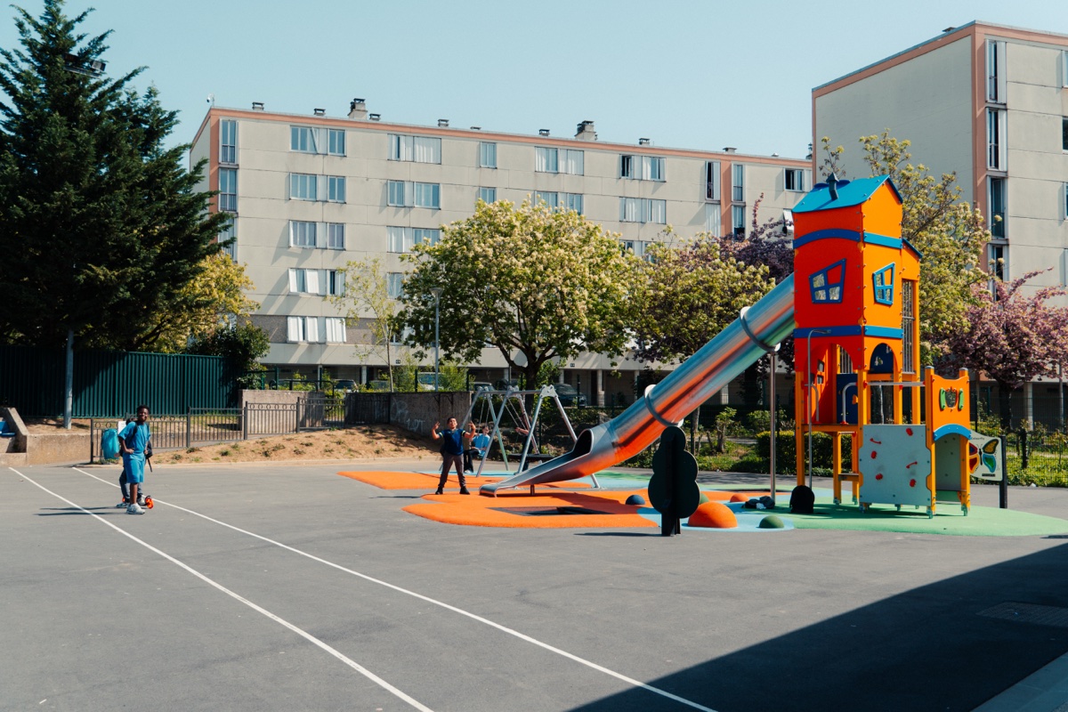 Aire de jeux urbaine avec structure et toboggan, sol EPDM Aire de jeux urbaine avec structure de jeux colorée orange et bleue, toboggan, sol EPDM orange, enfants en train de jouer dans un contexte de quartier résidentiel