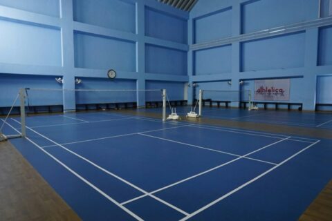Vue d'ensemble d'une salle de badminton avec deux terrains réglementaires côte à côte : sol bleu, lignes blanches, poteaux de filet et hauteur sous plafond