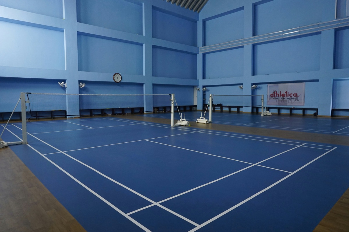 Salle de badminton, deux terrains réglementaires côte à côte Vue d'ensemble d'une salle de badminton avec deux terrains réglementaires côte à côte : sol bleu, lignes blanches, poteaux de filet et hauteur sous plafond