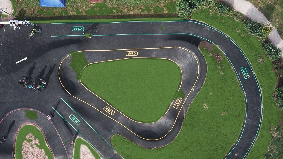 Vue de drone rapprochée d'un pumptrack en enrobé : marquages jaunes et verts de sens de circulation et de niveaux, riders au départ