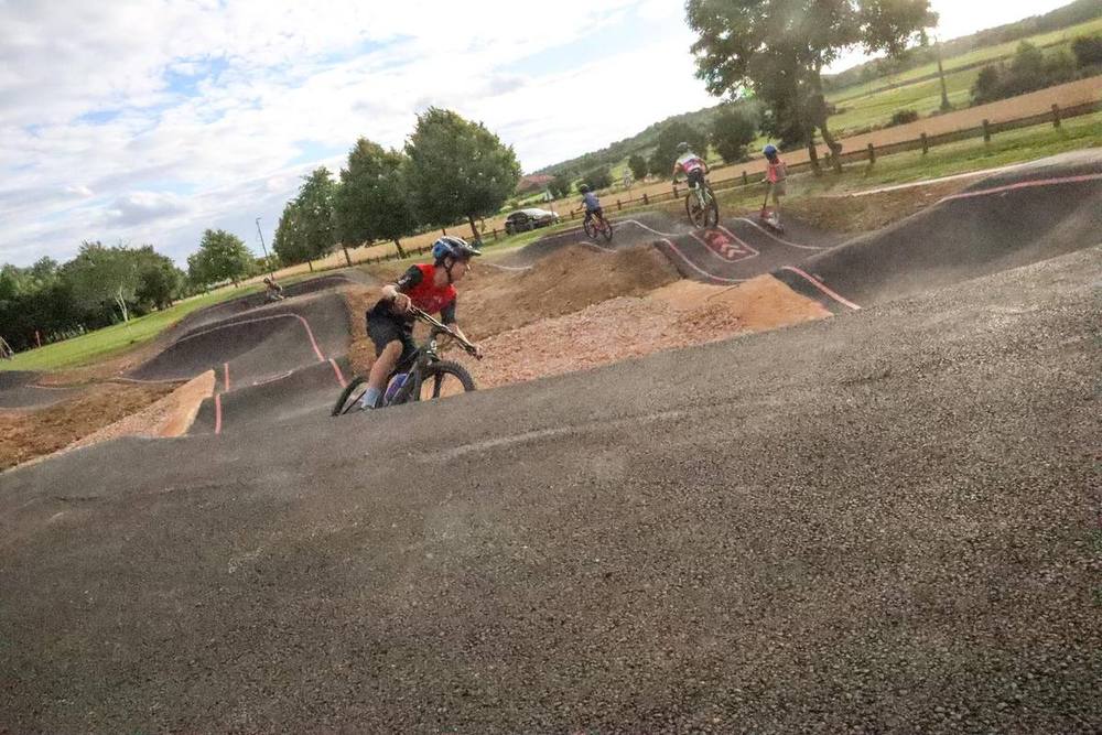 Jeune rider à VTT prenant un virage relevé sur un pumptrack en enrobé, d'autres pratiquants sur les bosses