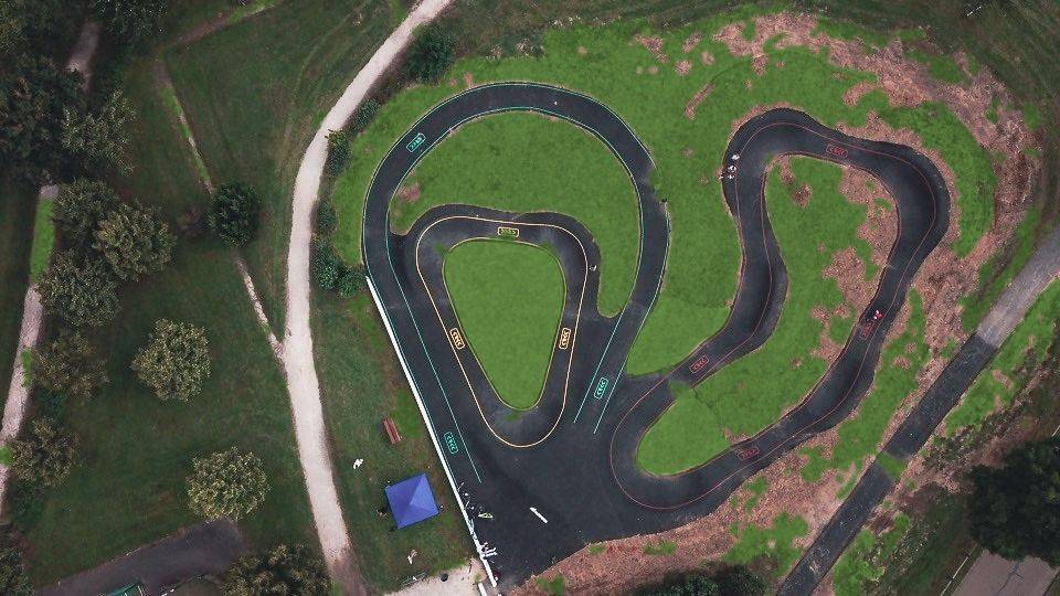 Vue zénithale d'un pumptrack en enrobé avec virages relevés, lignes de marquage colorées et îlots engazonnés